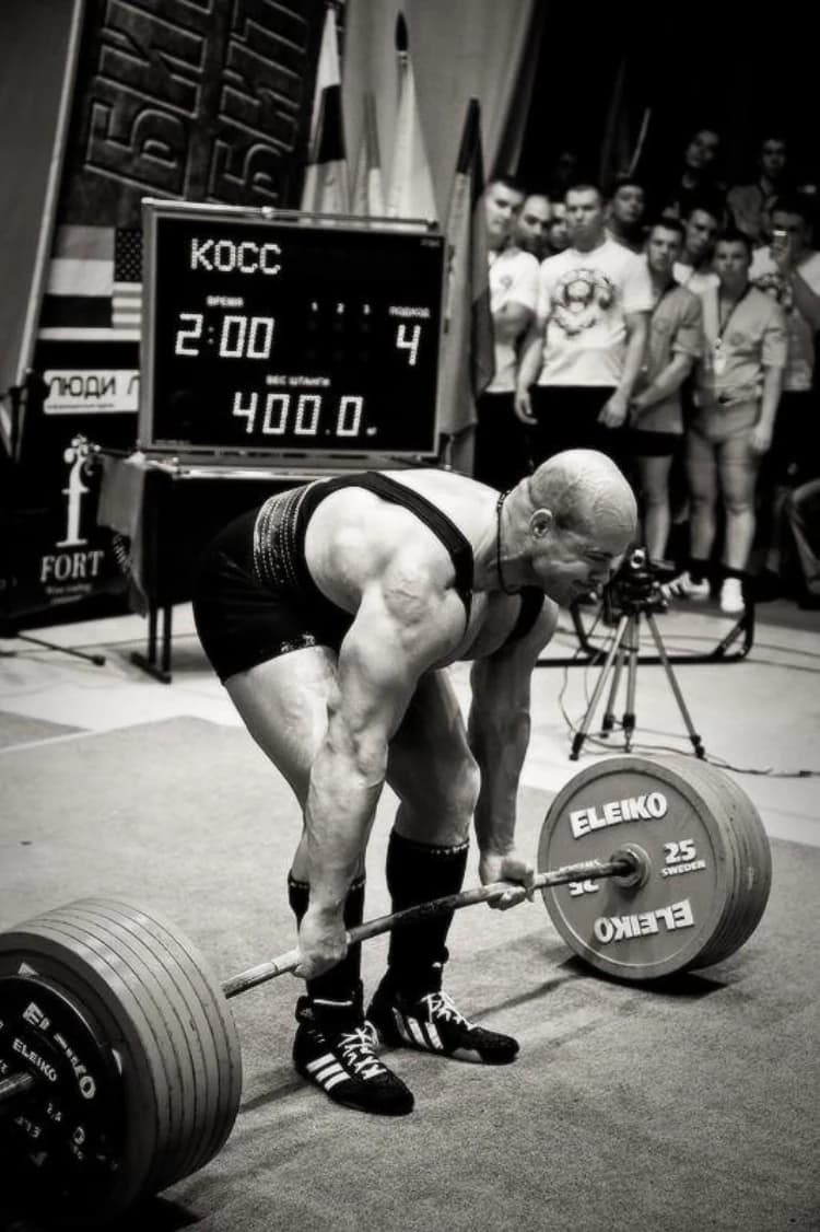 A lifter sets his grip over a loaded bar in black and white, the meet scoreboard behind him showing a 400.0 attempt beneath a row of national flags.